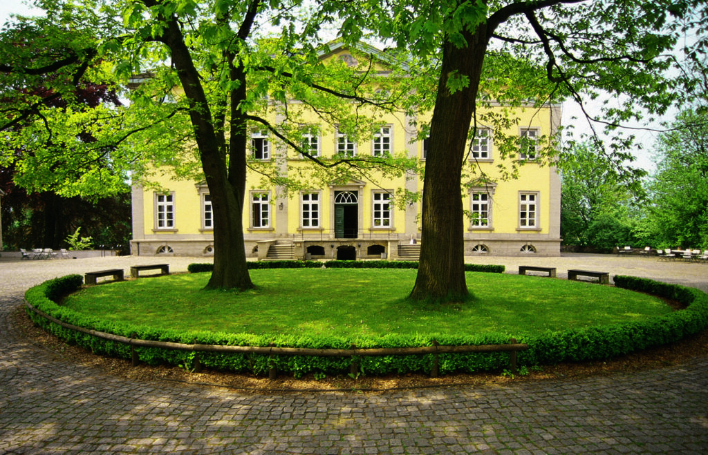 Gelbes Herrenhaus in Villigst, umgeben von altem Baumbestand und gepflastertem Hof mit runder Grünfläche.