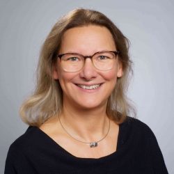 Porträt von Nicole Manns-Rodenbach, einer lächelnden Frau mit Brille und schulterlangem Haar vor neutralem Hintergrund.